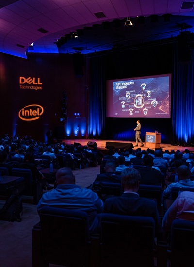 A room full of seated people watching a presentation given by a person on a stage with Dell and Intel signage