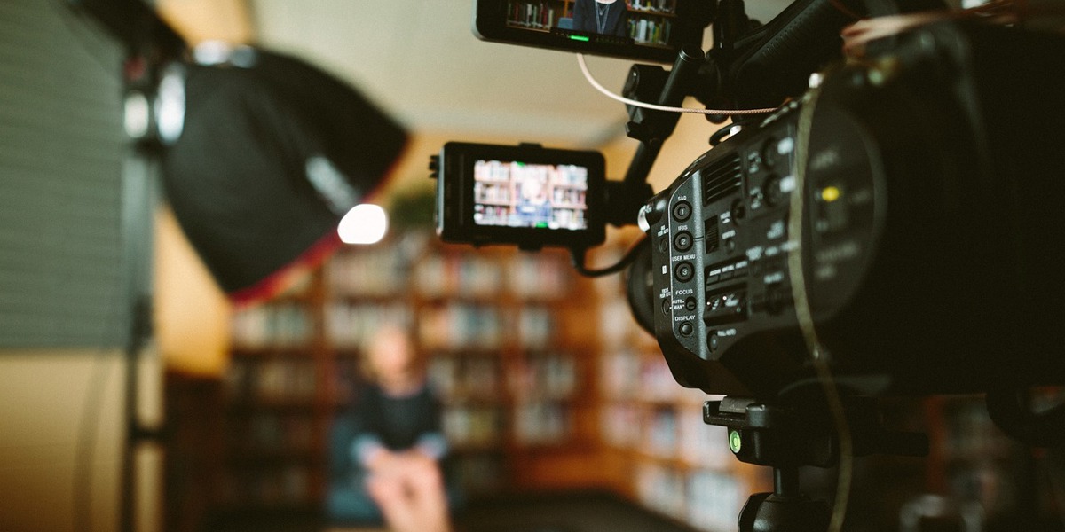 Close-up of a video camera recording a person sitting in front of a bookshelf with studio lighting.