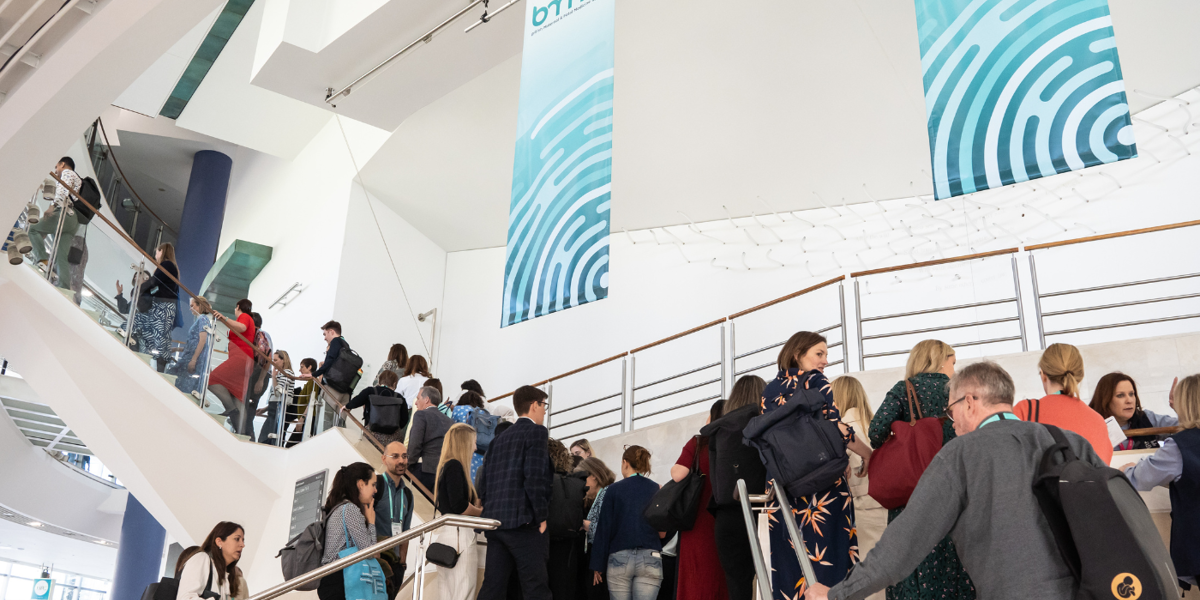 People on a staircase entering the BMFMS Annual Conference 2025 at the ICC Belfast with two large blue banners hanging 
