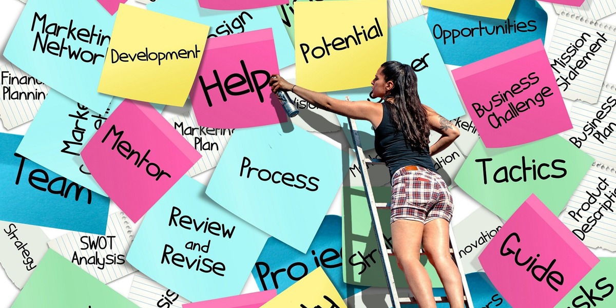 Woman on a ladder placing colourful post-it notes on a wall; notes display words like projects, process, team, mentor, and guide.