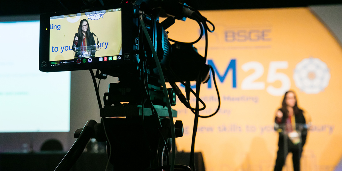 Video camera filming a conference speaker, with the speaker visible on the camera screen