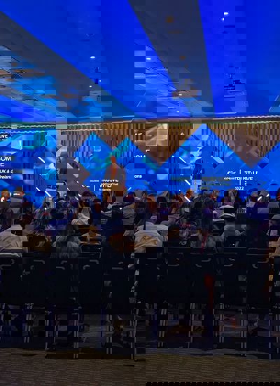 A room full of seated people watching a presentation given by a person on a stage