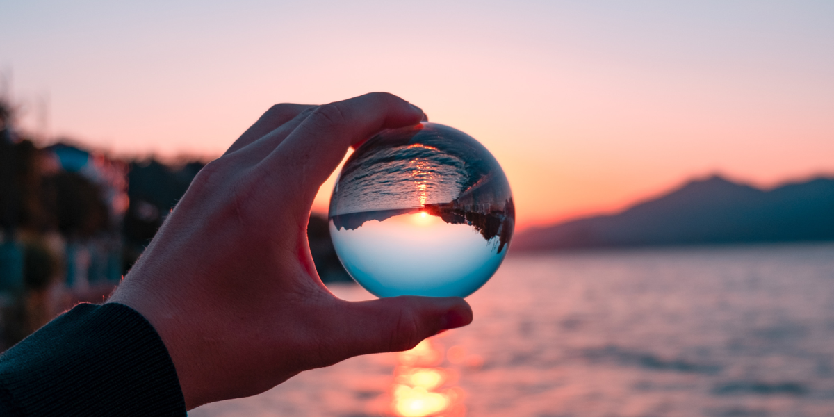 Person holding a glass ball with the sea and a sunset visible through the ball in the background.